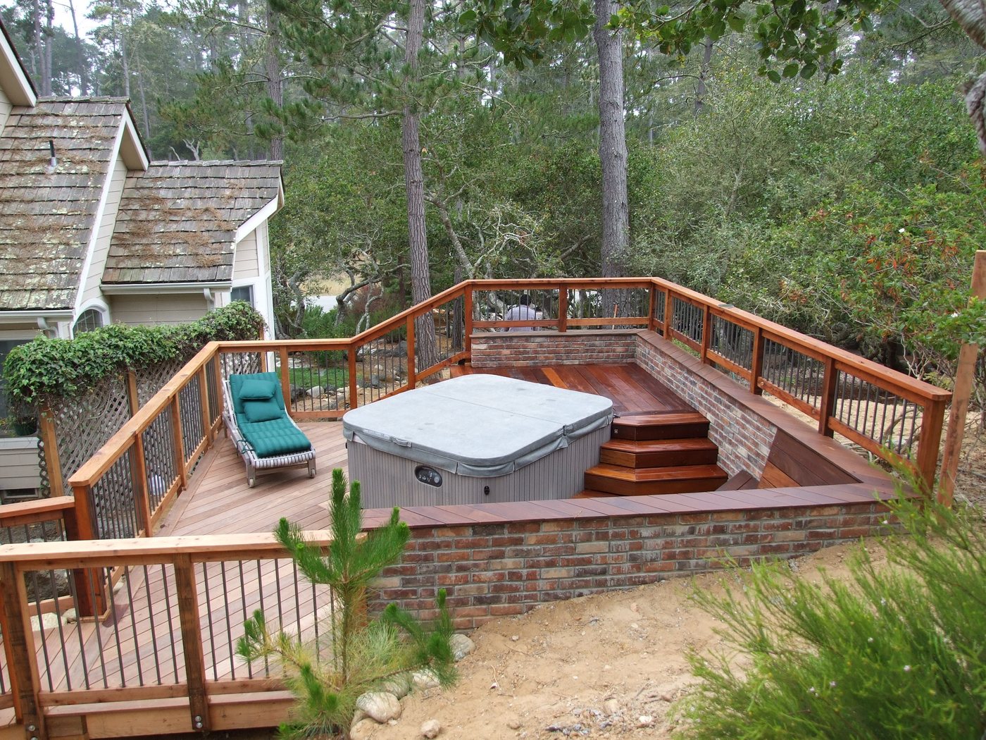 A hot tub set into a wood deck with a brick base, wood railings and a built-in step, among pine trees.