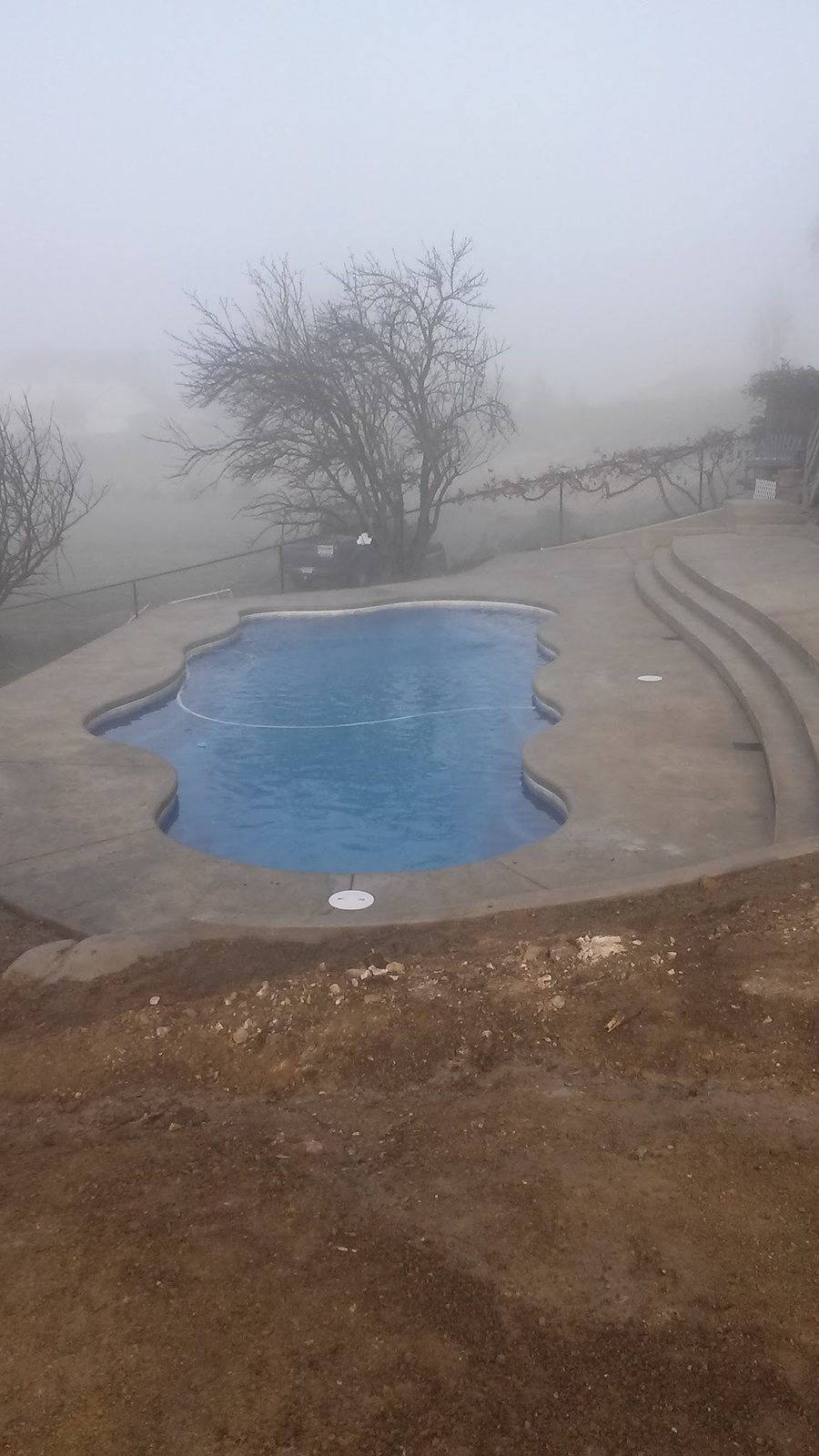 A pool with blue water and a stone-look deck on a hillside, in morning fog, with fresh dirt around it.