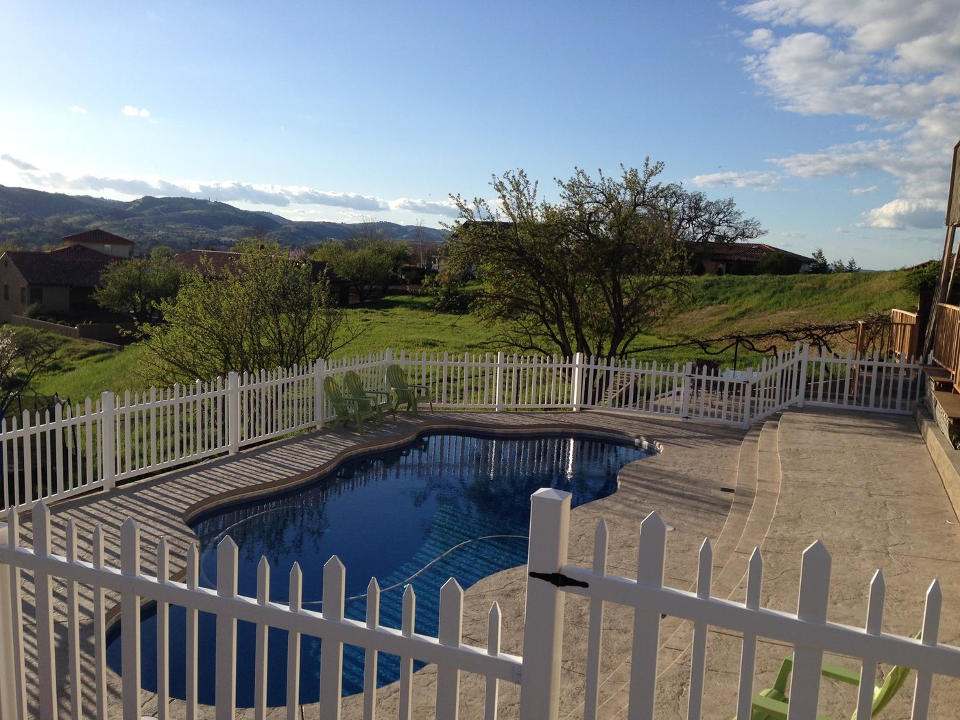 A kidney-shaped pool with dark blue water and a stone-look deck, behind a white picket fence, with green hills beyond.